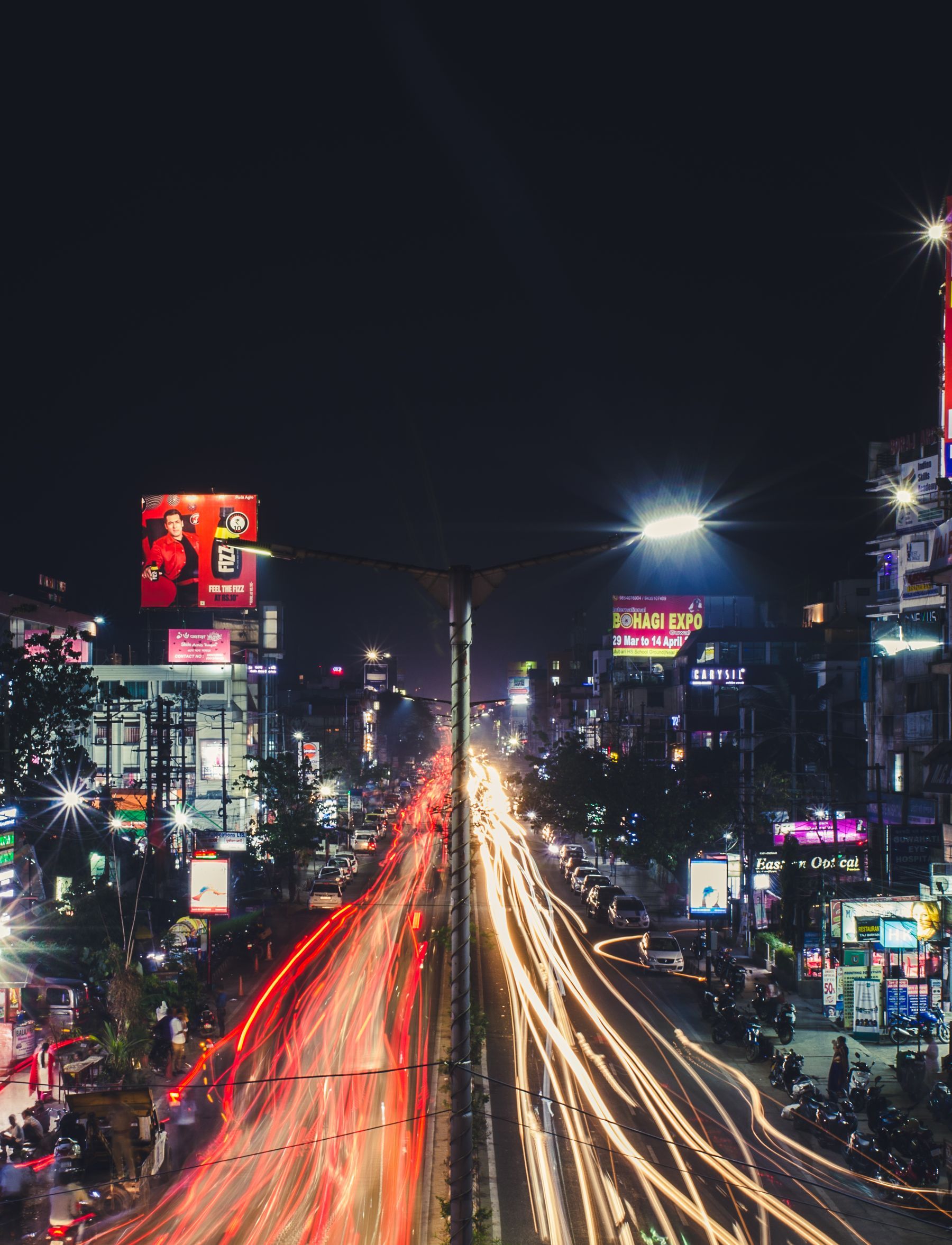 Hoardings lining a busy Indian road at night, traffic streaking past